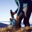 Person placing a black ESBIT SCULPTOR 1L vacuum flask into a blue backpack outdoors on a clear day