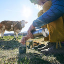 Person using ESBIT solid fuel stove and stainless steel pot outdoors with cow and mountains in background