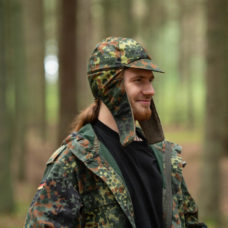 Man wearing genuine German Army flecktarn winter pile cap with ear flaps and matching camo jacket in forest