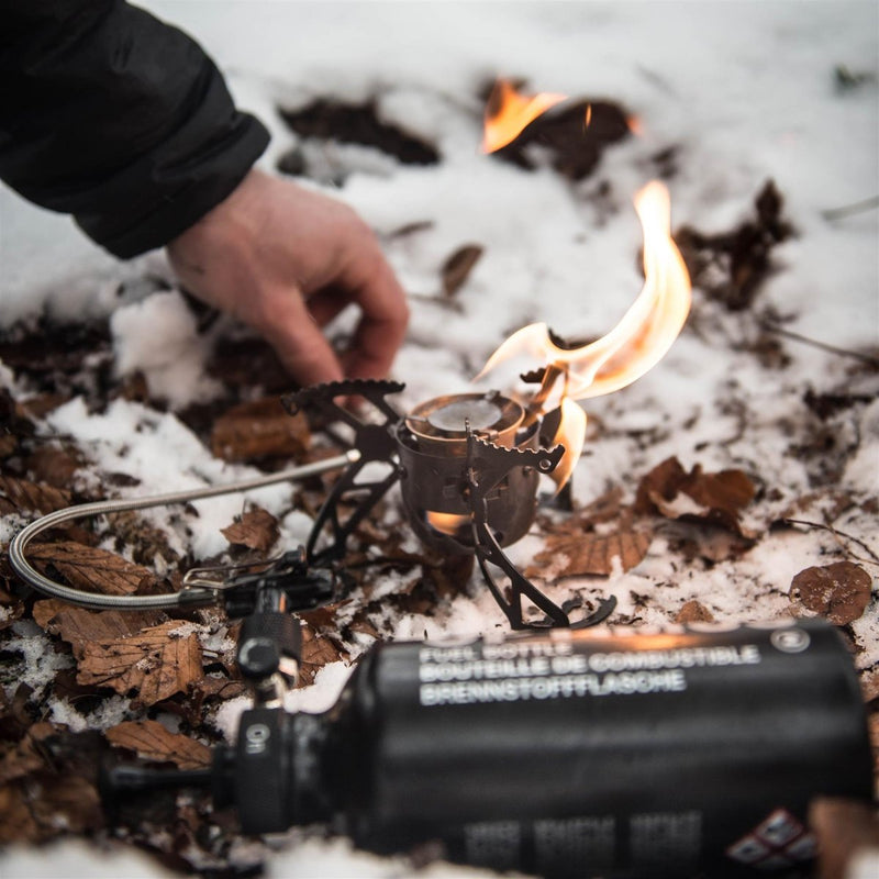 Optimus Polaris black camping stove with visible flame, used outdoors on snow and dry leaves with fuel bottle nearby