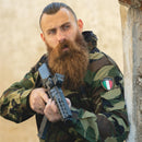 Man wearing Original Italy military combat jacket in woodland camo aiming firearm while leaning on wall outdoors.