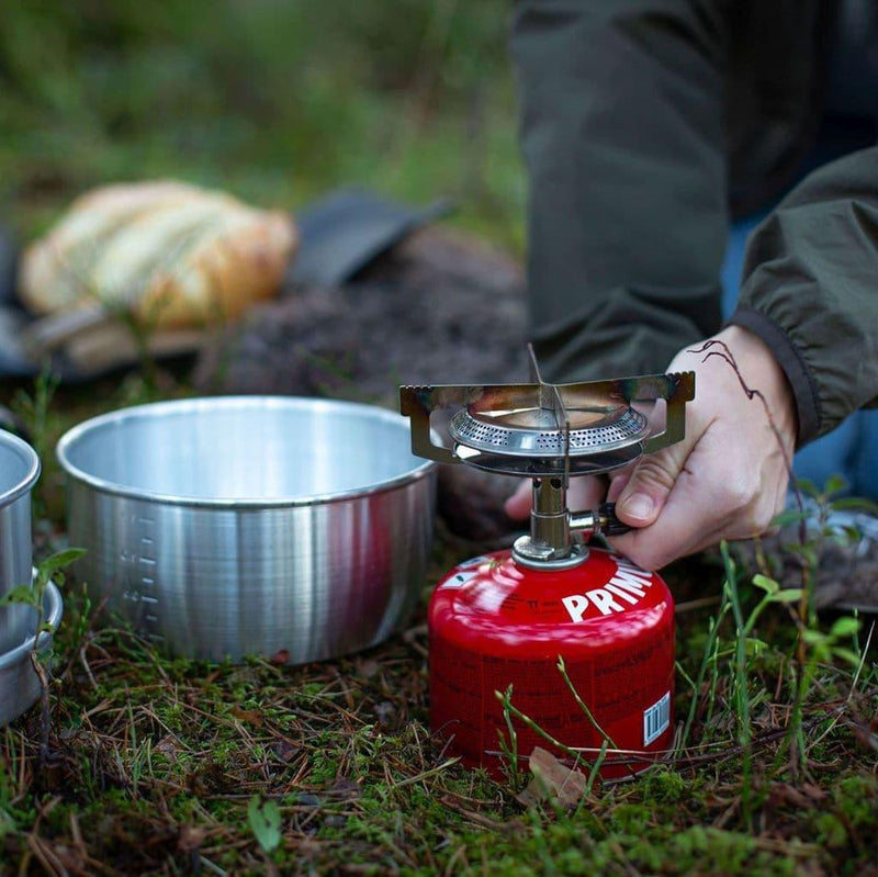 Person adjusting a compact red Primus Mimer propane camping stove outdoors next to a metal cooking pot on grass.